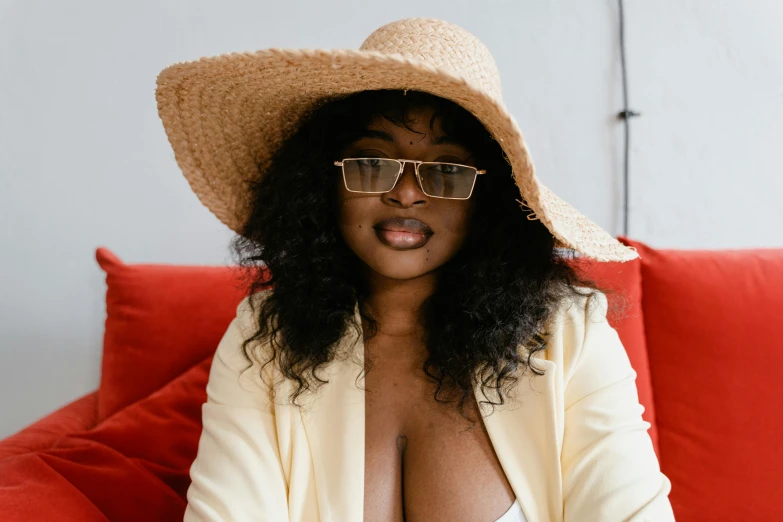 a woman wearing a hat and glasses sitting on a couch, by Carey Morris, trending on pexels, afrofuturism, buxom chested, square rimmed glasses, wearing wide sunhat, dressed in a robe