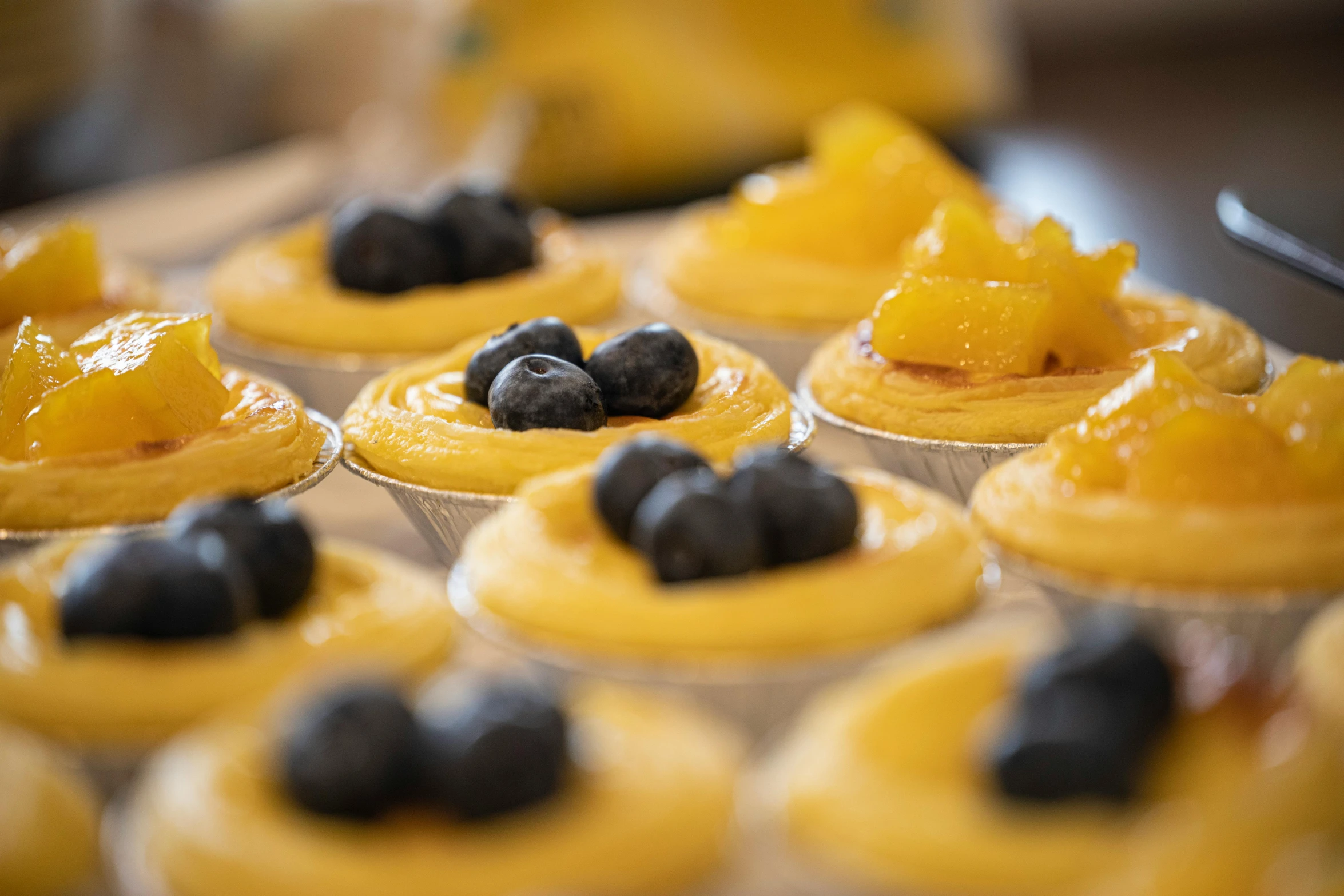 a close up of a tray of cupcakes with blueberries, by Lee Loughridge, unsplash, yellow colours, flan, thumbnail, brazilian