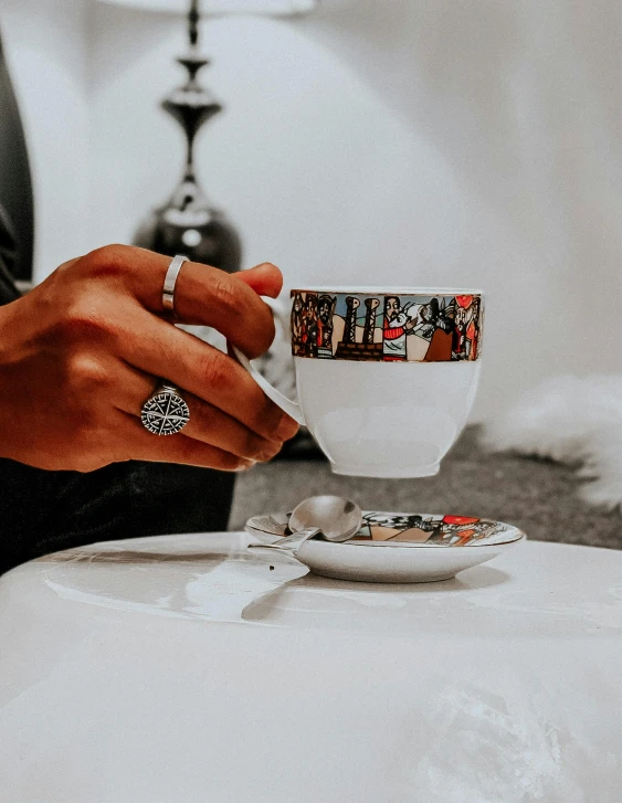 a person sitting at a table with a cup and saucer, by Lucia Peka, pexels contest winner, cloisonnism, silver white gold red details, middle eastern details, sleek hands, instagram story