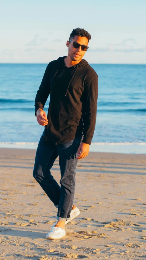 a man standing on a beach next to the ocean, wearing jeans and a black hoodie, main colour - black, long tail, hero shot