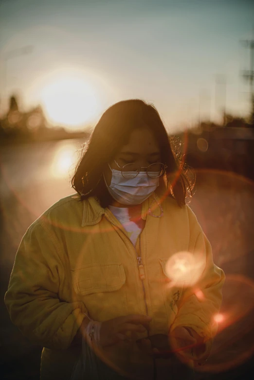 a woman wearing a face mask standing next to a body of water, a picture, trending on pexels, happening, yellow light, asian sun, healthcare worker, woman in streetwear