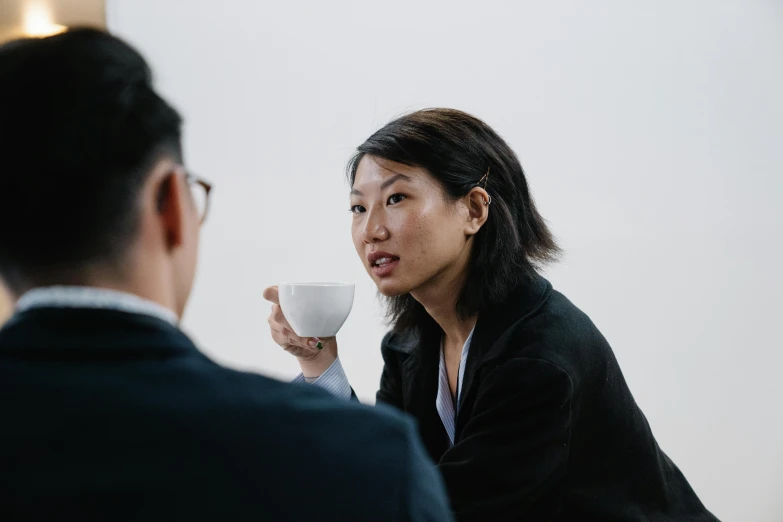 a woman sitting at a table with a cup of coffee, unsplash, zeen chin and farel dalrymple, woman in black business suit, wondering about others, asian male