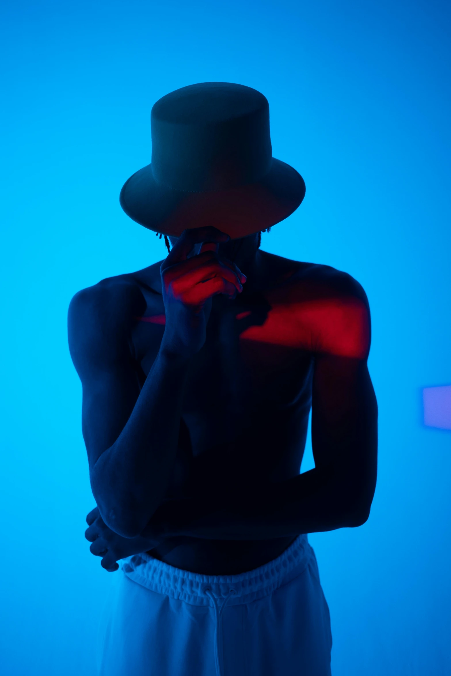 a man with a hat standing in front of a blue background, inspired by Carrie Mae Weems, conceptual art, colored gel lighting, lascivious pose, red light, (mist)