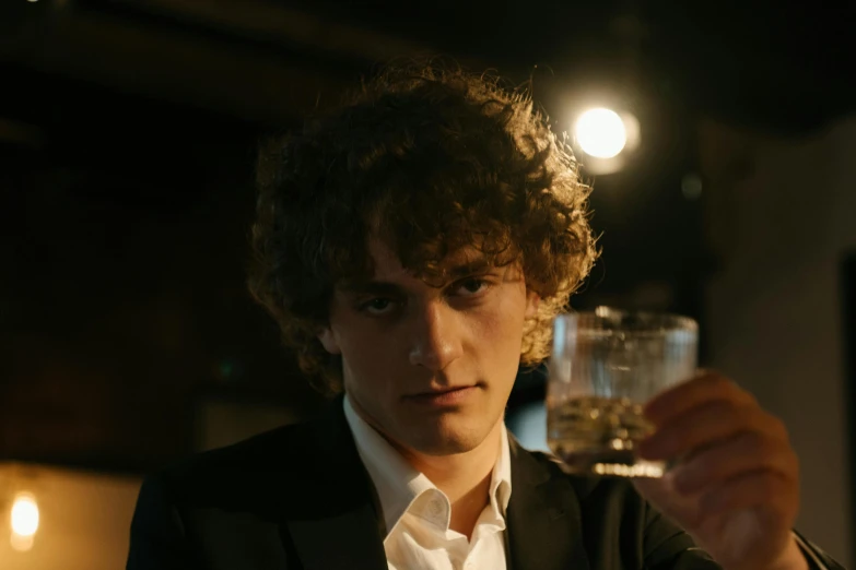 a man in a suit holding a glass of wine, an album cover, inspired by Nan Goldin, pexels contest winner, renaissance, timothee chalamet, curly haired, [ theatrical ], drinking a glass of whiskey