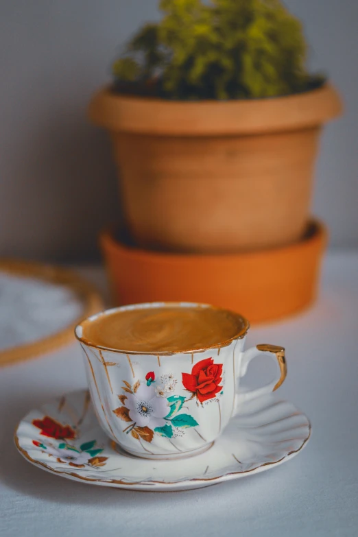 a cup of coffee on a saucer next to a potted plant, a still life, trending on pexels, art nouveau, middle eastern, brown flowers, colored milk tea, product shot