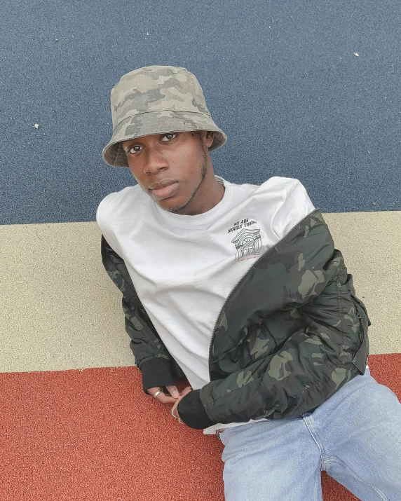 a man in a hat leaning against a wall, by Robbie Trevino, wearing a track suit, camouflage uniform, black teenage boy, subreddit / r / whale