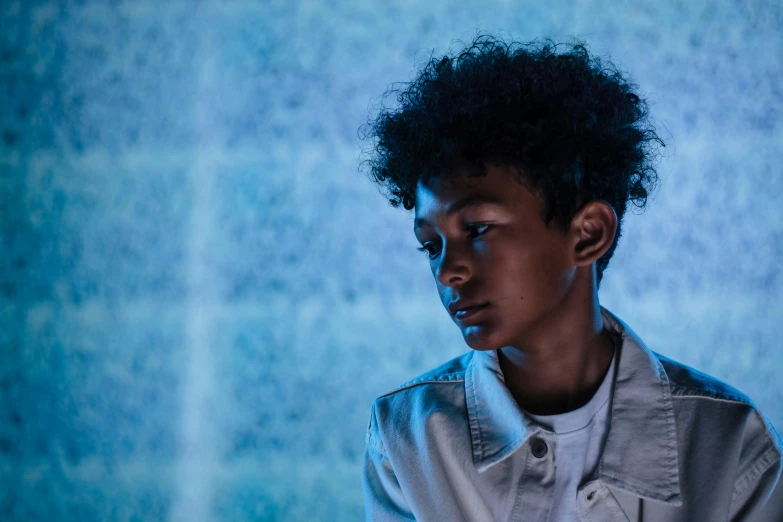 a young man standing in front of a blue wall, an album cover, by John Hutton, pexels contest winner, magical realism, willow smith young, dark moody backlighting, in the show westworld, finn wolfhard
