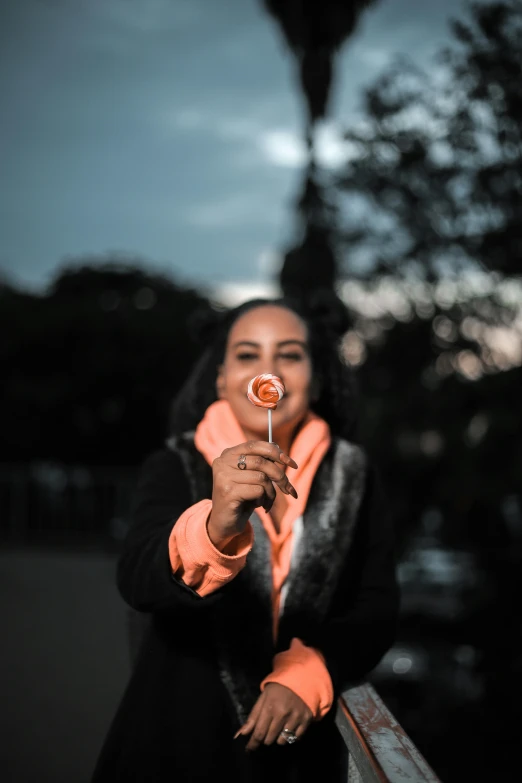 a woman holding a lollipop in front of her face, pexels contest winner, black and orange, early evening, ( ( dark skin ) ), in a city park