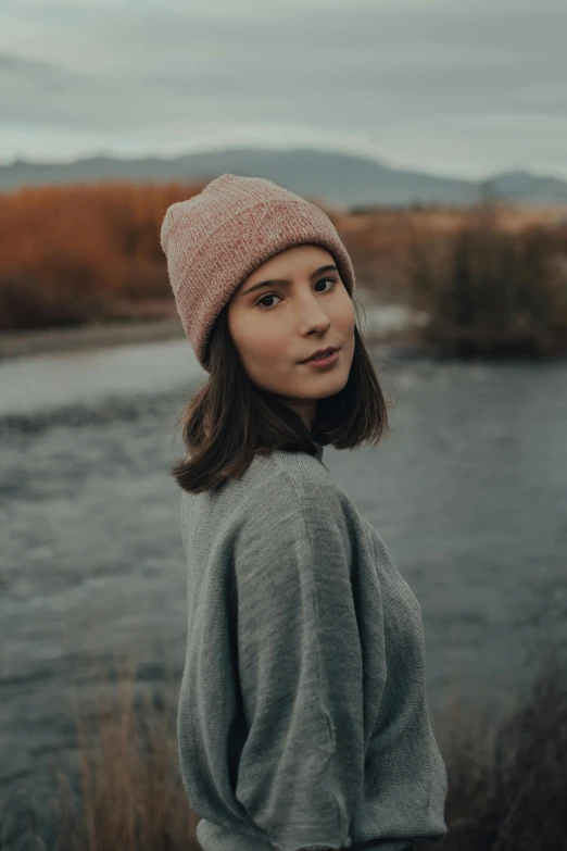 a woman standing in front of a river, wearing beanie, muted colors. ue 5, sweater, rosy cheeks