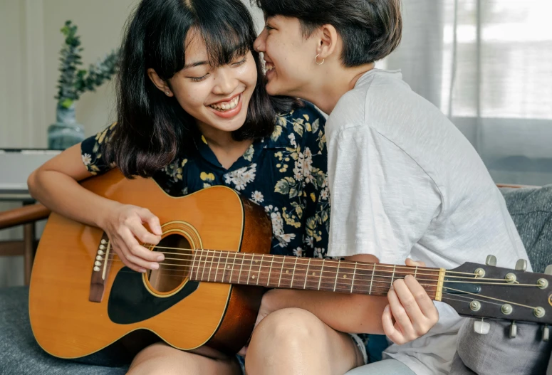 a man and a woman sitting on a couch playing a guitar, trending on pexels, lesbian embrace, an asian woman, smiling slightly, thumbnail