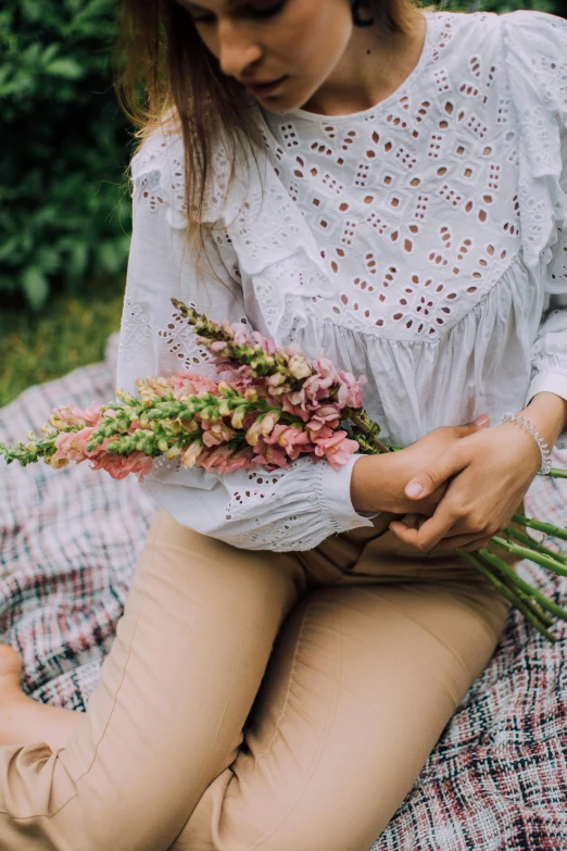 a woman sitting on a blanket holding a bunch of flowers, by Alice Mason, trending on unsplash, renaissance, wearing a white shirt, pale pink grass, detail shot, color image