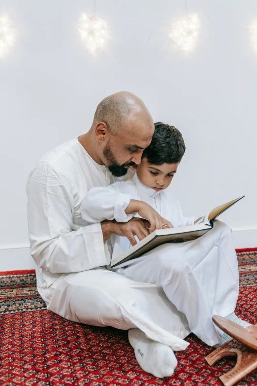 a man sitting on the floor reading a book to a young boy, hurufiyya, professionally done, religious, thumbnail, father figure image