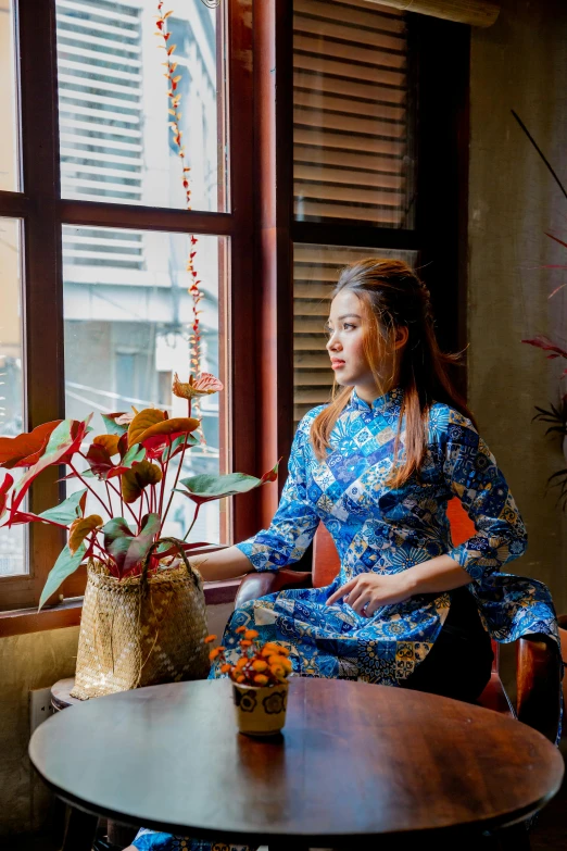 a woman sitting at a table in front of a window, inspired by Cui Bai, ao dai, blue, cosy atmosphere, square