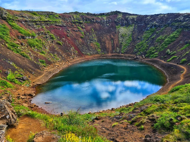 a large crater filled with water next to a lush green hillside, by Hallsteinn Sigurðsson, pexels contest winner, hurufiyya, blue and green and red tones, brown, flowers around, white
