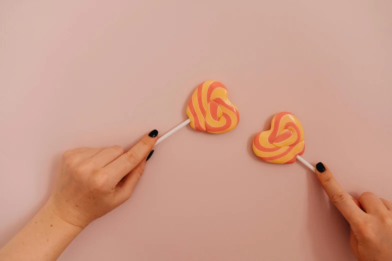 two hands holding two lollipops on a pink surface, inspired by Ödön Márffy, trending on pexels, swirls, caramel, carefully crafted, neon orange