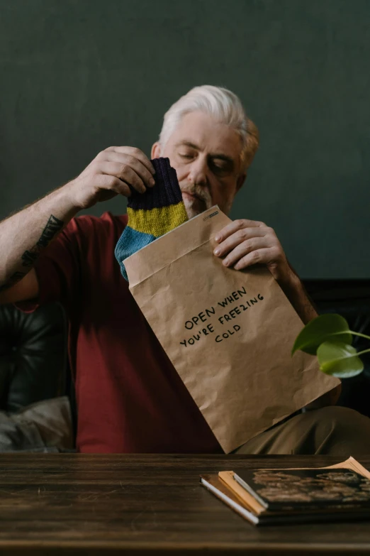 a man sitting on a couch holding a paper bag, a colorized photo, by Jesper Knudsen, pexels contest winner, playful vibe, packaging, older male, opening
