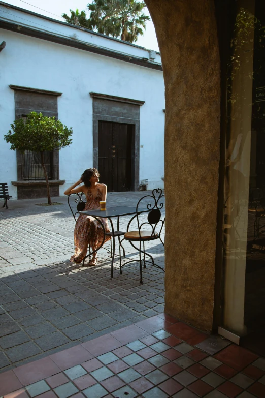 a woman sitting at a table in a courtyard, a portrait, inspired by Luis Paret y Alcazar, trending on unsplash, downtown mexico, morning coffee, distant full body view, full sun