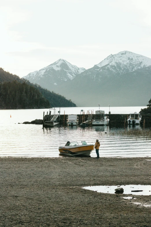 a man standing on top of a sandy beach next to a body of water, boat, patagonian, boat dock, multiple stories