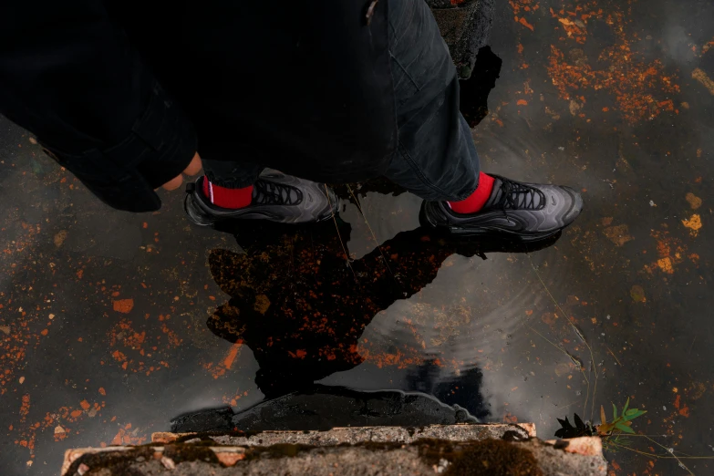 a person standing next to a puddle of water, black and red, sneakers, smouldering charred timber, looking down on the camera