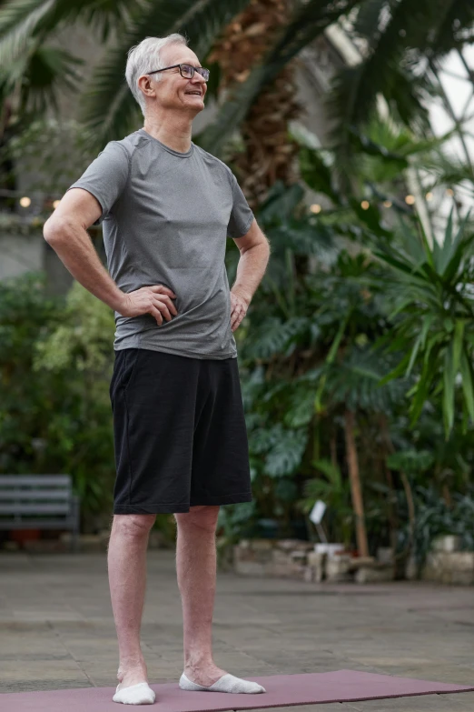 a man standing on a yoga mat with his hands on his hips, a portrait, shutterstock, standing in a botanical garden, gray shorts and black socks, wearing a black t-shirt, very tall and slender