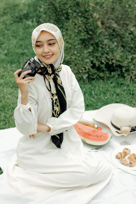 a woman sitting on a blanket holding a camera, a colorized photo, by Basuki Abdullah, trending on pexels, hurufiyya, wearing a melon, ( ivory black ), having a picnic, with