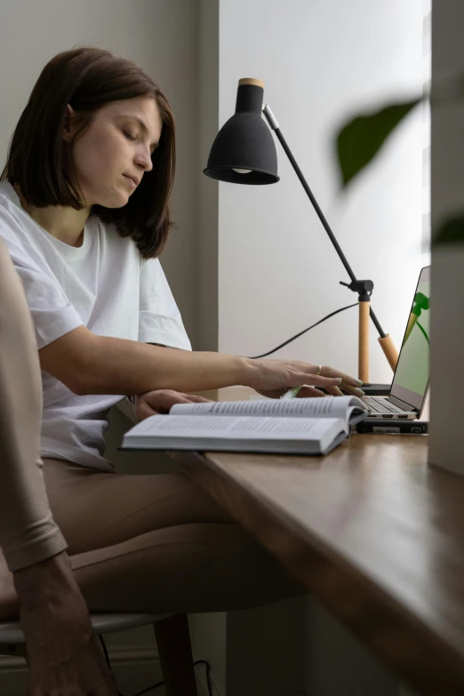 a woman sitting at a desk using a laptop computer, by Carey Morris, pexels contest winner, light academia aesthetic, profile image, holding a book, full body angle