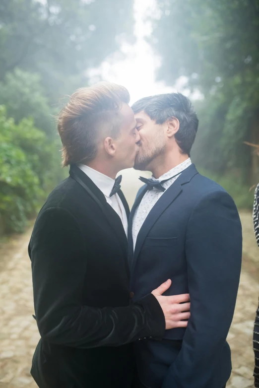 a couple of people that are kissing each other, two handsome men, well - dressed, lush surroundings, press shot