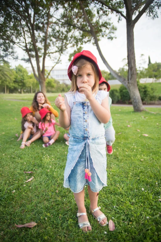 a little girl standing on top of a lush green field, an album cover, inspired by Elsa Beskow, pexels contest winner, barbecuing chewing gum, red hat, denim, in a city park