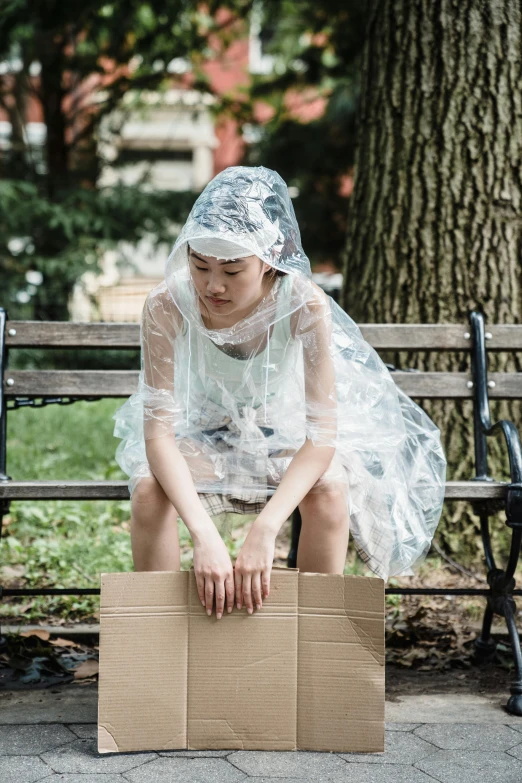 a woman sitting on a bench next to a cardboard box, by Ellen Gallagher, unsplash, process art, wearing a plastic garbage bag, white transparent veil, young asian woman, hailstorm