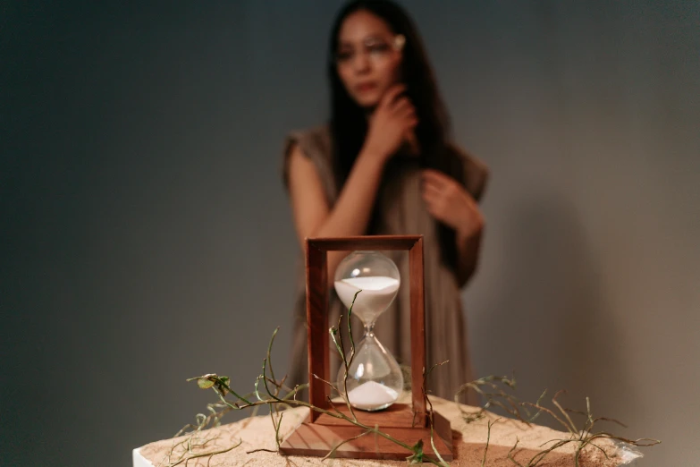 a woman standing next to a hourglass on a table, an album cover, inspired by Diego Giacometti, unsplash, kinetic art, a wooden, cinematic full shot, ignant, 3 / 4 view portrait