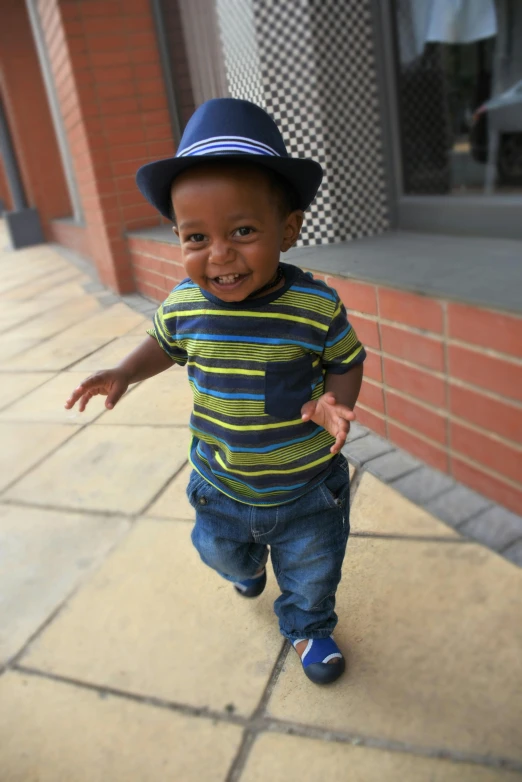 a little boy that is standing on a sidewalk, wearing a hat, running towards camera, riyahd cassiem, smiling playfully