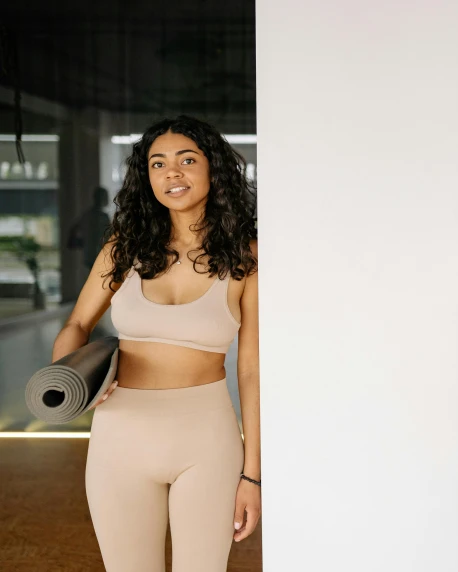 a woman standing next to a wall holding a yoga mat, happening, with brown skin, wearing bra, trending photo, candid portrait photo