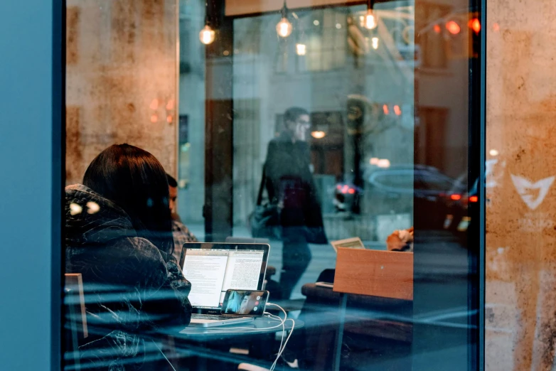 a person sitting at a table with a laptop, by Emma Andijewska, pexels contest winner, happening, shopwindows, background bar, looking through frosted glass, some people are sitting
