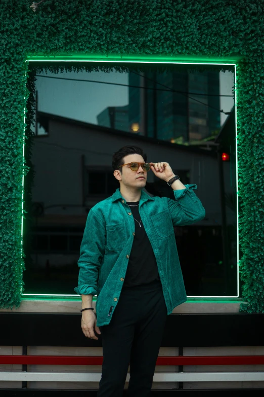 a man in a green jacket standing in front of a building, an album cover, by Alejandro Obregón, pexels contest winner, zachary quinto, khyzyl saleem, nightlife, man with glasses