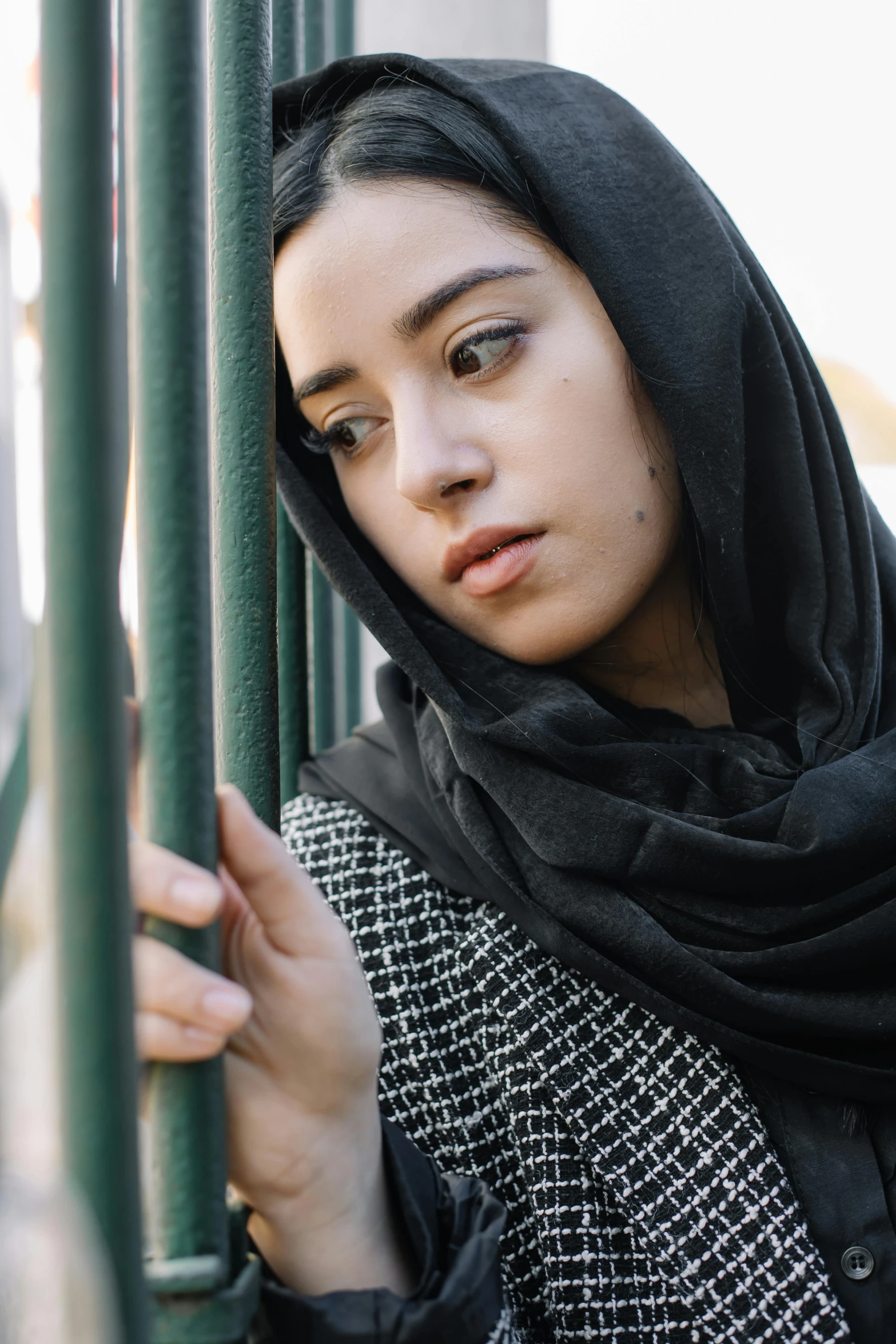 a woman in a black and white scarf leaning against a pole, inspired by Maryam Hashemi, trending on unsplash, standing astride a gate, concerned expression, young woman's face, square