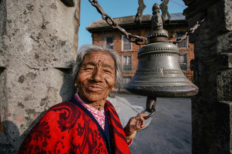 a woman holding a bell in front of a building, inspired by Steve McCurry, pexels contest winner, cloisonnism, nepal, an elderly, 🦩🪐🐞👩🏻🦳, welcoming grin