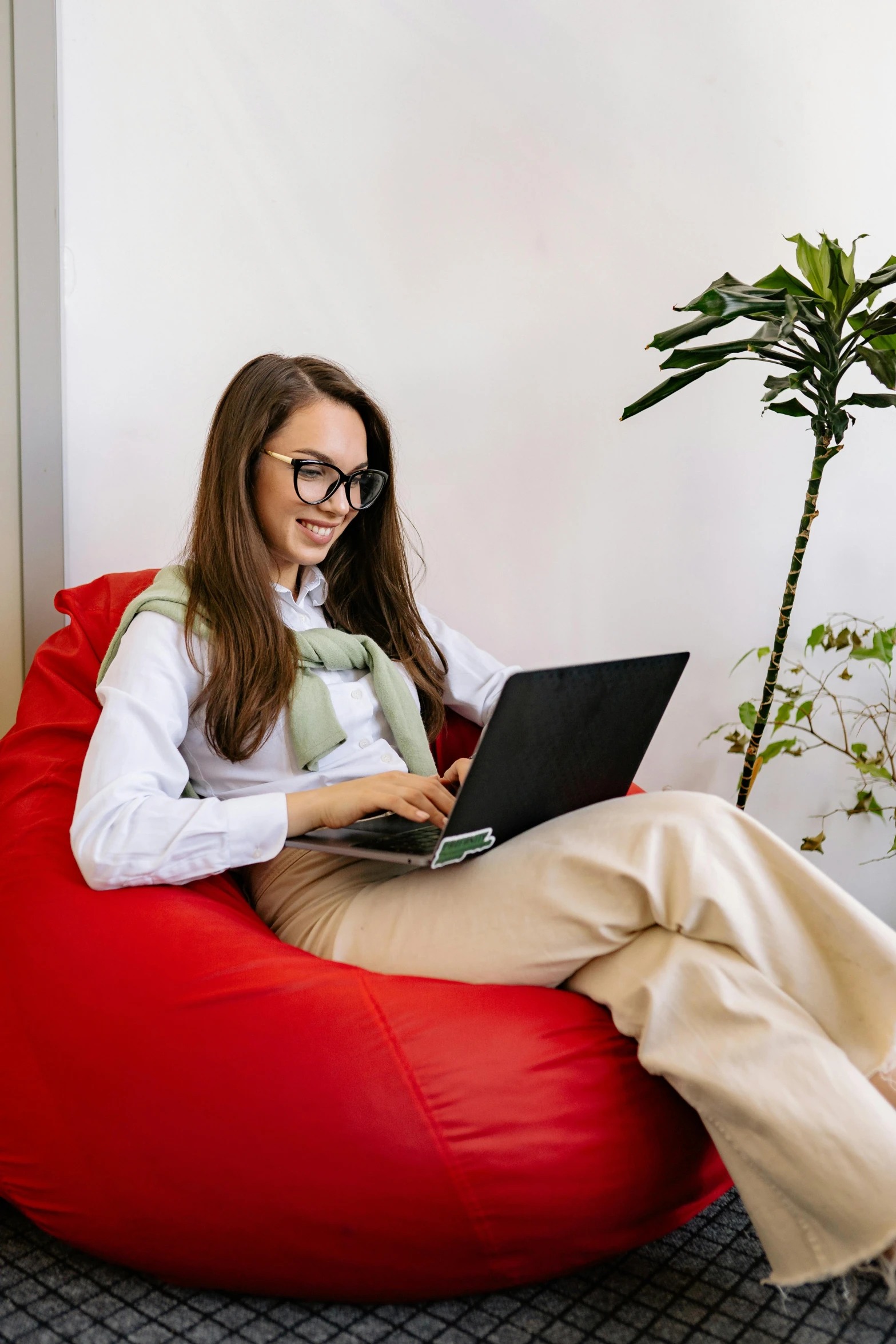 a woman sitting on a bean bag chair using a laptop, next to a plant, wearing small round glasses, sitting on a red button, hip corporate