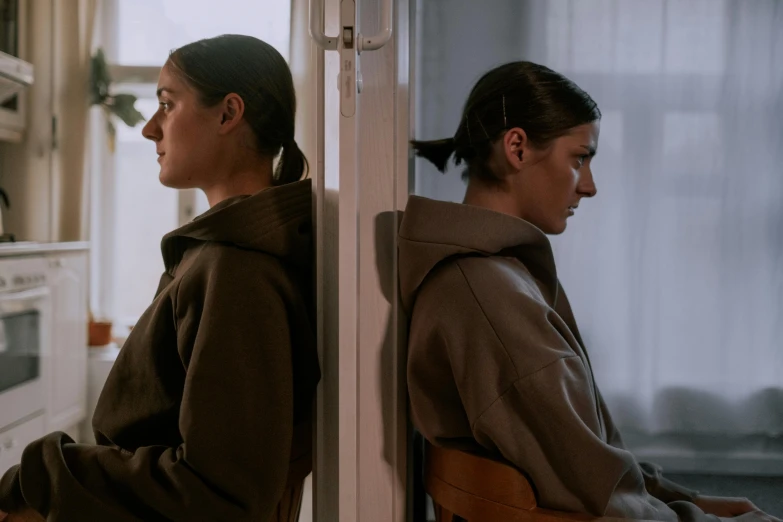 a couple of women sitting next to each other in a kitchen, by Emma Andijewska, purism, wearing brown robes, mirrored, still from a live action movie, hunched shoulders