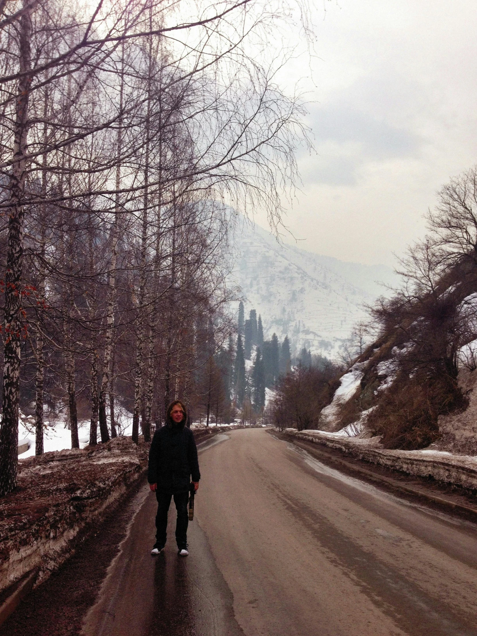 a man riding a skateboard down a snow covered road, inspired by Fyodor Vasilyev, standing in front of a mountain, with dark trees in foreground, taken in the late 2000s, album photo