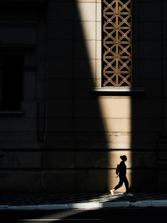 a man walking down a street past a tall building, unsplash contest winner, light and space, female image in shadow, holy light, photographed for reuters, ( ray of sunlight )