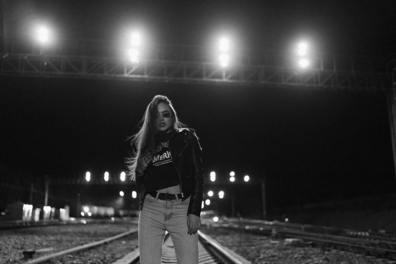 a woman standing on a train track at night, a black and white photo, unsplash, female streetwear blogger, concert photo, she wears leather jacket, ariel perez