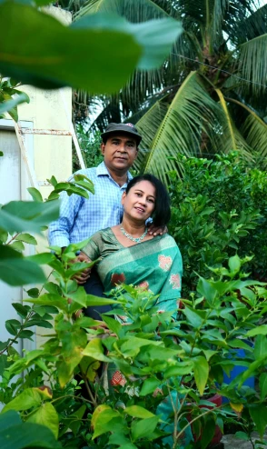 a man standing next to a woman in a green sari, large plants in the background, portrait image