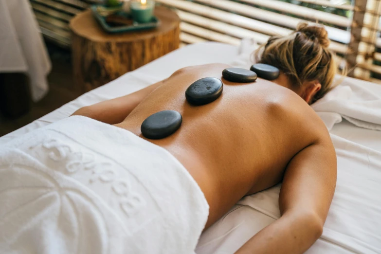 a woman laying on a bed with hot stones on her back, trending on pexels, animation, lush botany, grey robes, on a wooden table