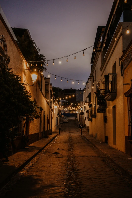 a street filled with lots of lights next to tall buildings, a portrait, by Alejandro Obregón, trending on unsplash, renaissance, tlaquepaque, the city of santa barbara, colonial era street, cafe lighting