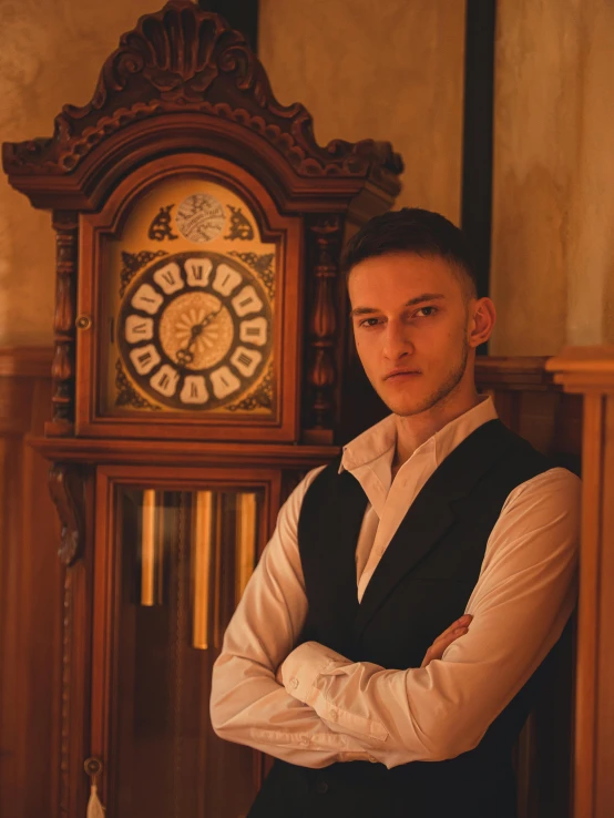 a man in a vest standing in front of a wooden grandfather clock