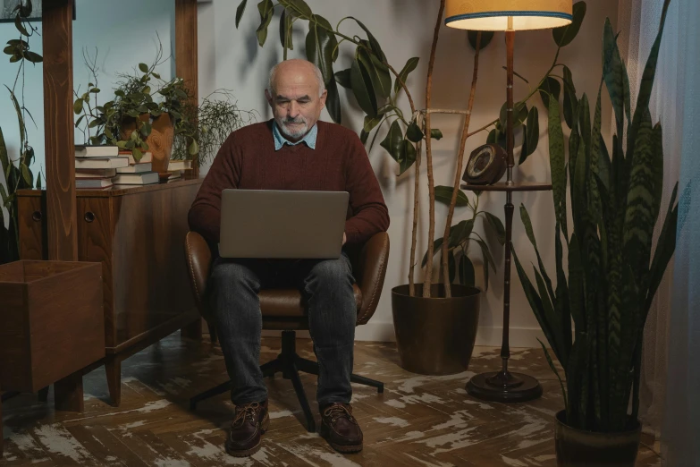 an old man is using his laptop while in a room with potted plants