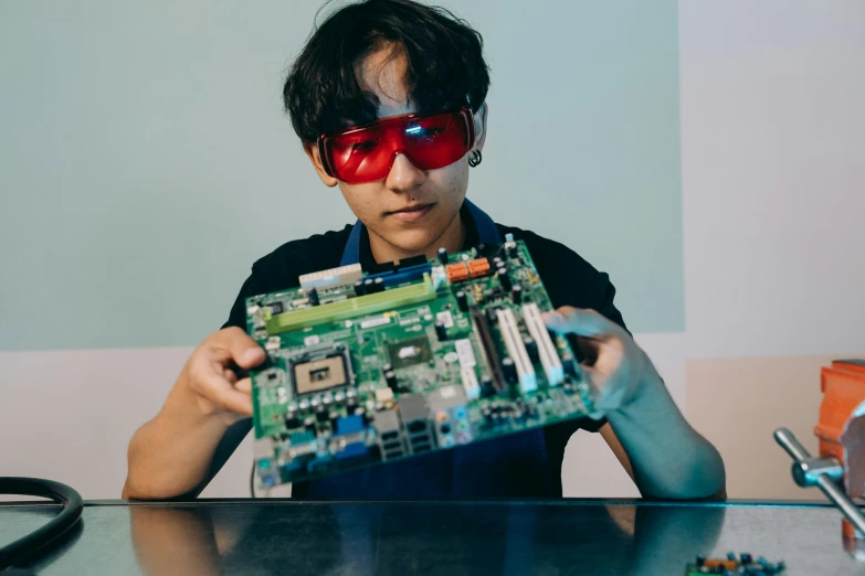 a man sitting at a table holding a computer motherboard, by Byron Galvez, unsplash, interactive art, wearing goggles, red laser, high school, an epic non - binary model