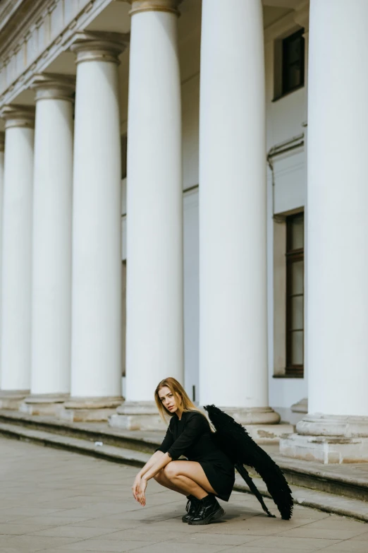 a woman kneeling on the ground in front of a building, by Tamas Galambos, pexels contest winner, neoclassicism, wearing a fancy black jacket, britt marling style, in front of white back drop, colonnade