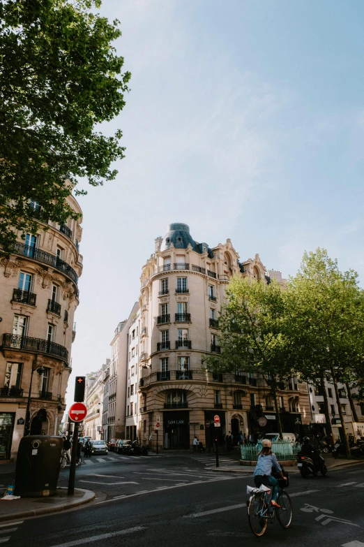a man riding a bike down a street next to tall buildings, trending on unsplash, paris school, trees around, rounded architecture, french village exterior, clear blue skies
