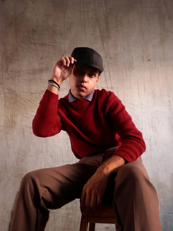 a man sitting on top of a wooden chair, an album cover, inspired by Alejandro Obregón, instagram, realism, wearing newsboy cap, red sweater and gray pants, depressed dramatic bicep pose, headshot profile picture